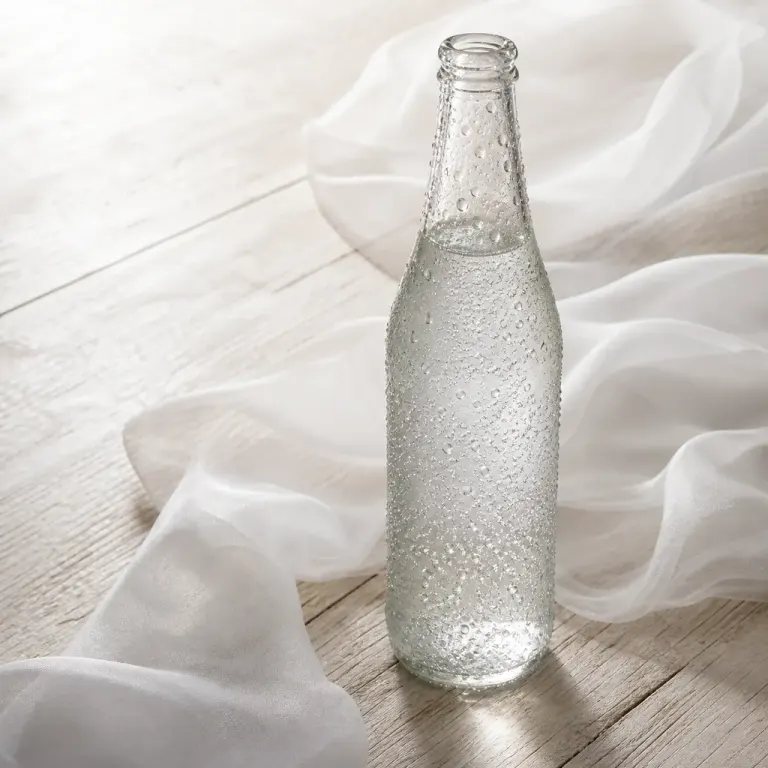 Clear carbonated beverage bottle with condensation glowing in backlight on bleached wood and silk